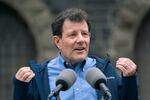 Nicholas Kristof wears a blue button down shirt and a darker blue jacket and gestures with his hands while he stands at a mirophone.