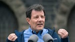 Nicholas Kristof wears a blue button down shirt and a darker blue jacket and gestures with his hands while he stands at a mirophone.