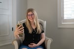Kaleigh Brendle sits in a beige rocking chair while holding a smartphone in front of her body. She's looking at the phone and smiling.