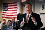 Mike Pompeo holds a microphone in his right hand while he speaks to a room of people with an American flag on the wall in the background. Maskless people are visible in the background.