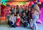 A group of drag performers and their supporters pose for a picture in front of a colorful backdrop