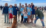 Six adults and four kids pose smiling in the sand, with their dog on a leash. The kids are wearing their first place medals.