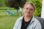 Amber Petersen sits in the backyard of her parents’ home in Iowa City, Iowa. She has blonde hair pulled back and is wearing glasses. Her husband and children are playing in a hammock behind her.