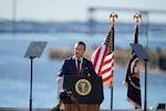 Standing outdoors and behind a lectern with a microphone attached to it, Defense Secretary Pete Hegseth speaks at a Maryland shipyard this week. A body of water is in the background.