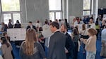 A crowd of people stand in a room with posters displayed on easels with a science fair feel to it.