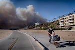 A man pushes his mother-in-law in a shopping cart as smoke fills the sky in the distance behind them. 