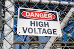 Undated photo of a "Danger: High Voltage" sign on chain link fence with power generators in the background at an unspecified location.