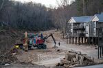 A precarious-looking wooden apartment complex is shown on stilts, on ground where a river flooded and receded. Big construction equipment is shown in front of the building.