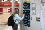 A high school student with long blonde hair wearing a black backpack places her cellphone in to a phone holder hanging on a wall as she enters class.