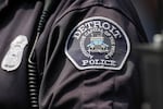 This photo shows a close-up of a police officer's uniform. A patch on the dark sleeve says: "Detroit Police" and "Automotive Capital of the World."
