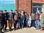 A smiling group of actors and crew members stand shoulder to shoulder in front of a brick building with the Oregon Film Trail placard in the center.