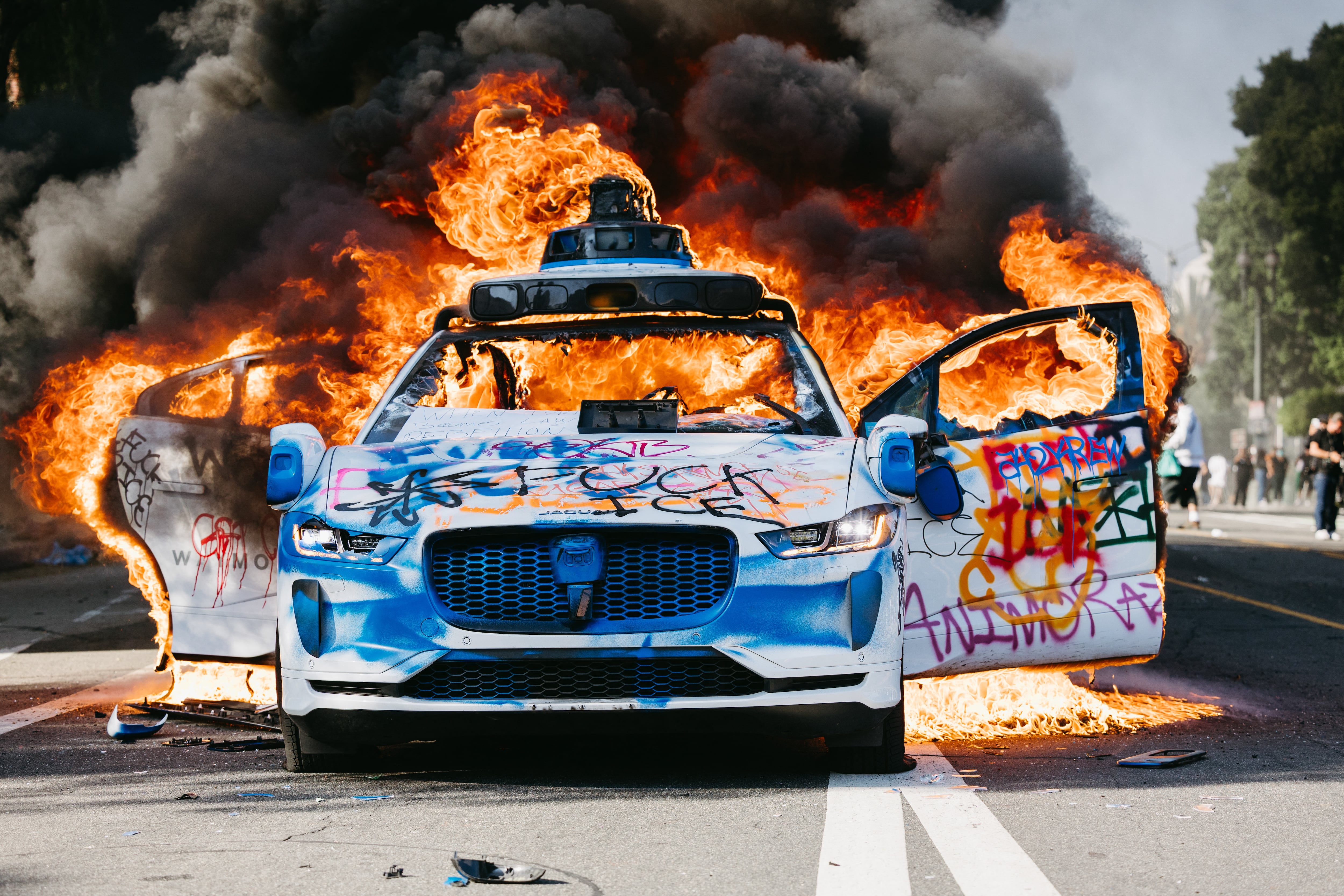 A Waymo car burns in the street as smoke billows during protests in Los Angeles on Sunday. Following a series of aggressive federal immigration operations in the city, tensions escalated when ICE agents conducted a high-profile raid on a Home Depot location.