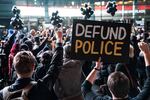 A large crowd of protesters is seen from behind. One person holds a sign that says "Defund police."