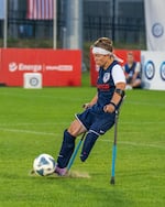 A woman on forearm crutches kicks a soccer ball with her right foot.