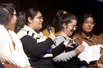 A woman in glasses and a white and black striped talk holds a microphone on stage. She is flanked by three other women who are listening intently.