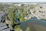 A river, seen from above, flows into a dam, and then emerges as a small creek. A row of houses overlooks the river on the left side, and a highway runs alongside it on the right side.