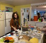 A person stands at a kitchen table surrounded by pies, vegetables and dishes of food.