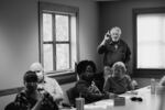 This black-and-white photo shows Ted Brown standing while talking about his past as a schoolteacher. He's wearing jeans and a button-down shirt. In front of him is a conference table at which other participants of the political discussion group are seated.