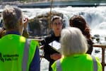 A woman in the foreground speaks to people in green safety jackets at the edge of the Willamette River with the falls in the background.