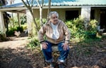 In this 2014 photo, Uruguayan President José Mujica sits outdoors on a chair while wearing blue jeans, a white button-down shirt and a partially unzipped light brown jacket. Behind him is his one-story home and a white chicken.