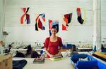 Britt Howard, the founder and owner of the Portland Garment Factory, pictured in her workspace.  She's standing with folded fabrics on a big table. Behind and around her are tools and sewing materials, some at smaller workstations in the background. Brightly colored fabric art hangs on a white wall behind her like flags.