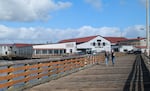 A pair of people walk down a wooden sidewalk at Astoria's Pier 39 on a sunny fall day. Behind them stands the Rogue Ales & Spirits building.