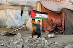 In this photo, two boys, one holding a Palestinian flag, run past tents at a camp for people displaced by conflict in Gaza. One tent is made from a white tarp displaying UNICEF's light blue logo. Concrete-looking rubble is piled on the ground.