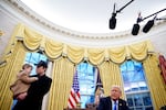 In this photo, taken at the Oval Office, President Trump is seated, and Elon Musk stands on the left side of the photo while holding his young son X Musk. Windows framed by yellow curtains are in the background.