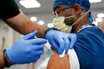 A person wearing medical scrubs and a face mask receives a shot in the upper arm from a person wearing medical gloves.