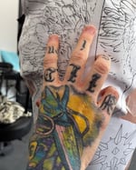 Close-up of a right hand, with a colorful tattoo on the back of the hand and text on the knuckles fingers, taping a piece of paper with an eagle design onto a person's leg.