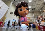 A team of people, including one on a tall ladder, pump up the  Dora the Explorer balloon inside a massive warehouse.