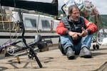 A man sits near a bike with a bag of recyclables on a wood dock near a boat