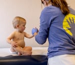 A child, who appears to be about a year old, is sitting on an exam table, wearing a diaper. A medical professional is holding the child by the hand.