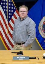 Mike Lorenson, who is both the mayor of Thief River Falls and an IT manager at DigiKey, poses for a photo at City Hall. He's standing behind a desk that has a gavel on it, as well as a nameplate that has Lorenson's name on it. He's wearing glasses and a long-sleeved, button-down shirt with a white, burgundy and dark green plaid-like pattern on it.