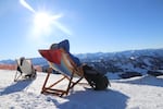 Two people sit on foldable lawn chairs that are set outdoors on snowy ground. In front of them is a vista of snowy mountains.