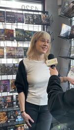 A woman with blond hair and glasses stands in the new releases section of a video store, smiling. Shelves full of DVDs to rent are behind her and to her left. She's being interviewed, and an arm holding an OPB microphone is partially in view.