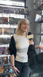 A woman with blond hair and glasses stands in the new releases section of a video store, smiling. Shelves full of DVDs to rent are behind her and to her left. She's being interviewed, and an arm holding an OPB microphone is partially in view.