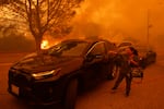 A woman stands outside a car with flames in the background and smoke all around. 