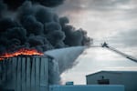 From a ladder, a firefighter douses flames with black smoke on the roof of a potato chip factory.