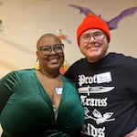 Two people stand side-by-side smiling into the camera. On the left is Board President Nocturne Webster in a green top and on the right is board treasurer Sami Webster in a black t-shirt that says "protect trans kids".