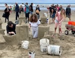 A man, woman and child kneel and stand on the beach in their competition plot, building a sandcastle together. The man is using a smoothing tool while the child uses a rake. Big white buckets and a smaller bucket for tools are in the foreground. The plot is roped off with red tape, and spectators are watching in the background.