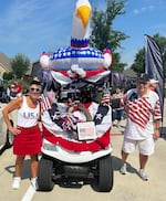 A woman, right, and a man, left, wearing patriotic American outfits pose with a Fourth of July decorated golf cart between them.