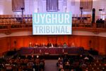 People sit in a large gallery facing a panel of other people. Above them all, a sign reads "Uyghur Tribunal"