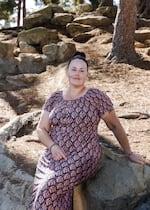 In this photo, Lisa Collenbaugh is sitting on a large rock outdoors. She's wearing a long dress with short sleeves that's black with a pink and tan pattern on it.