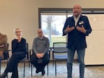 Greg Smith gestures at his chest in a conference room as he speaks. Kimberly Lindsay and Rick Stokoe sit quietly as they sit in a row of chairs set behind him.