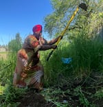 An African woman in a bright patterned dress and a red headwrap is seen in an action shot with a garden hoe held up high. She's on the edge of a patch of dirt with tall grasses in the background on a sunny day.