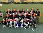 Pictured are 12 men in Black hockey jerseys. They form two lines, 6 sitting and 6 standing behind them. They have red trimming, and the men wear ice skates, black, and padding. One in the middle has a hockey stick, another on the far end does as well. On the far right side is one man without a jersey, he wears a black button up shirt with orange sleeves, dark brown pants.