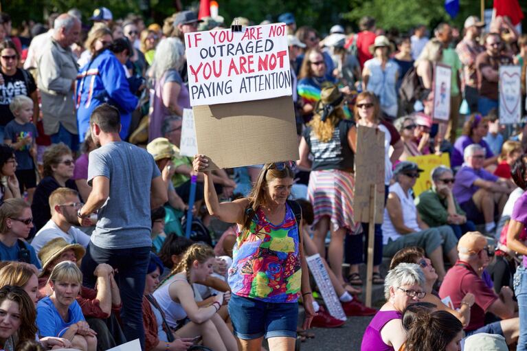 A woman carries a sign reading "If you are not outraged you are not paying attention" as she walks through a crowd of hundreds gathered at the Salmon Street Springs Fountain in Portland.