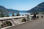 A cyclist stands on a bridge overlooking a river and hills.