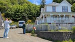 On a sunny day, two people stand by the famous "Goonies House" in Astoria. One holds a camera while the other poses for the picture with the house behind them.